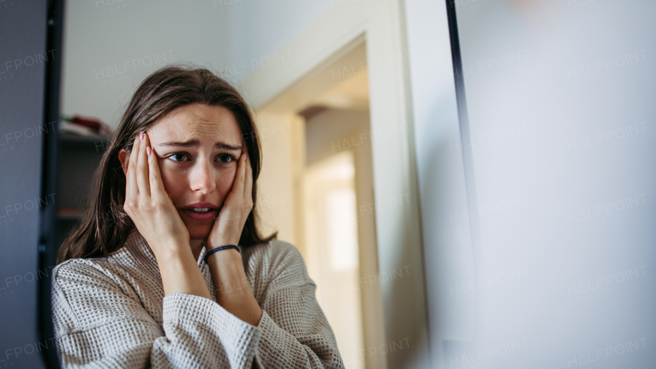 Sad depressed young woman alone at home, looking in mirror, feeling unhappy, lonely and anxious. Woman health problems and mental health.