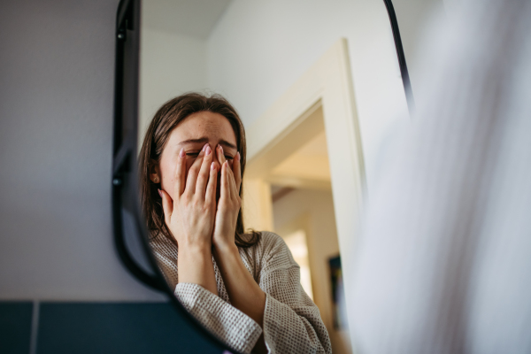 Sad depressed young woman alone at home, looking in mirror, feeling unhappy, lonely and anxious. Woman health problems and mental health.