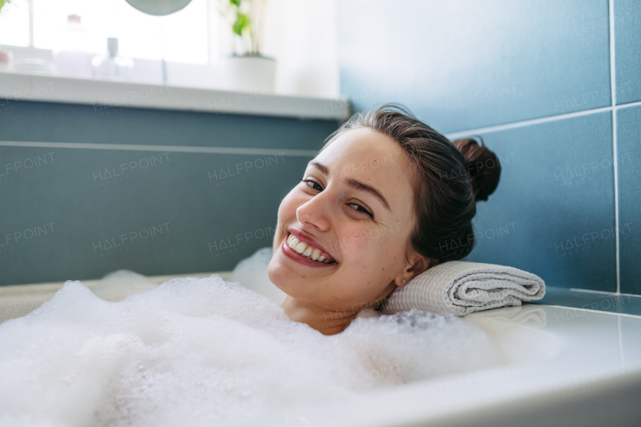 Beautiful woman relaxing in warm bathwater, soaking in bathtub. Enjoying bubble bath, smiling, looking at camera.