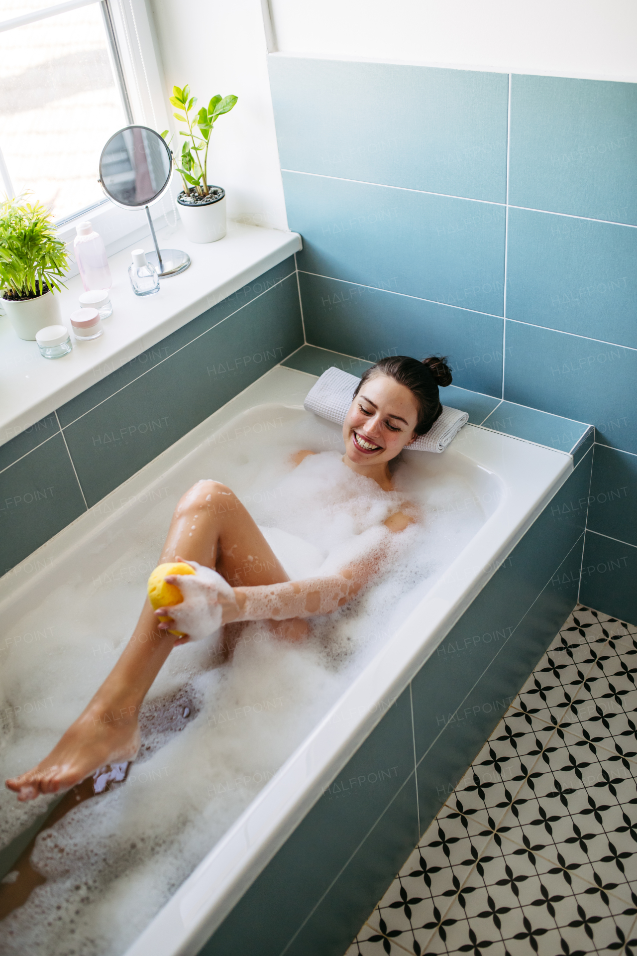 Top view of beautiful woman relaxing in warm bathwater, washing her body with sponge. Soaking in bathtub, enjoying bubble bath, smiling.