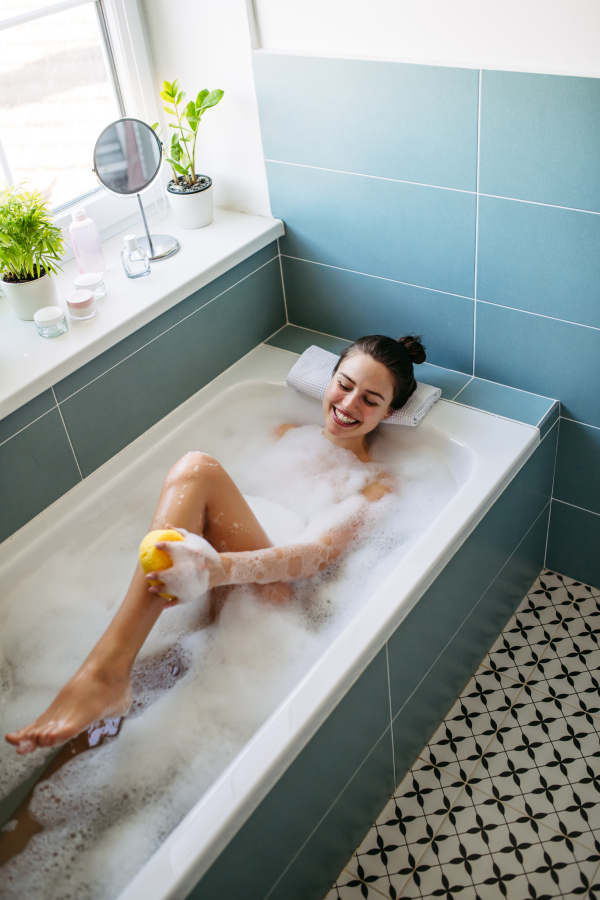 Top view of beautiful woman relaxing in warm bathwater, washing her body with sponge. Soaking in bathtub, enjoying bubble bath, smiling.