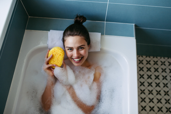 Top view of beautiful woman relaxing in warm bathwater, washing her body with sponge. Soaking in bathtub, enjoying bubble bath, smiling.