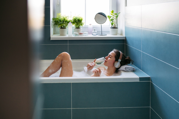 Beautiful woman relaxing in bathtub, listening music through headphones and drinking champagne, wine. Enjoying hot water in bubble bath with music.