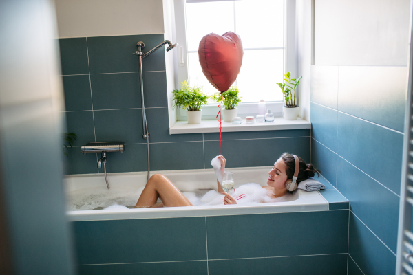 Beautiful woman relaxing in bathtub, listening music through headphones and drinking champagne, wine. Enjoying hot water in bubble bath with music. Holding heart shaped balloon.