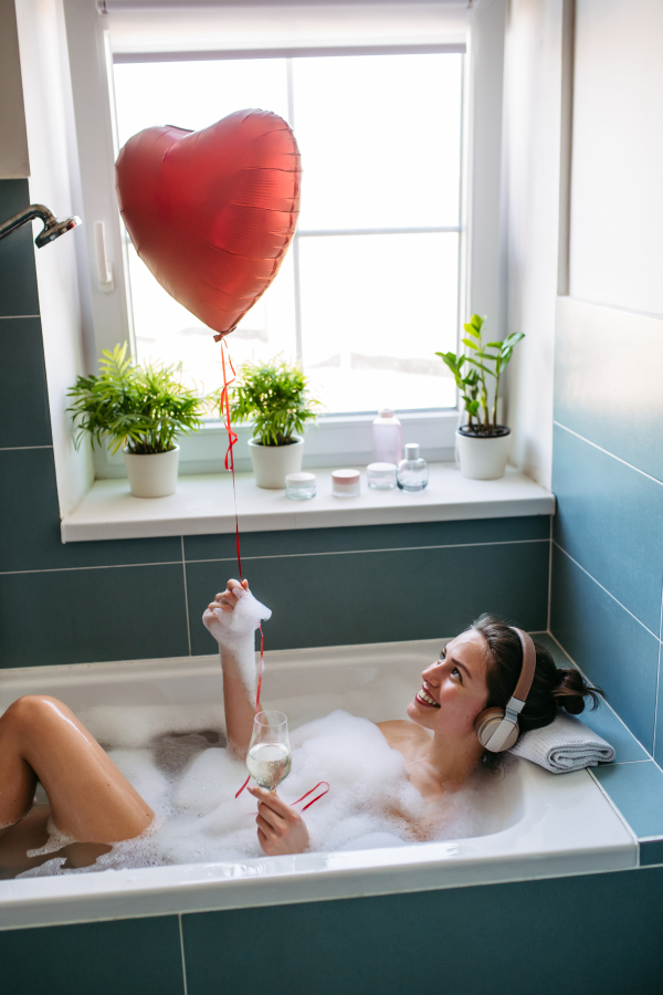 Beautiful woman relaxing in bathtub, listening music through headphones and drinking champagne, wine. Enjoying hot water in bubble bath with music. Holding heart shaped balloon.