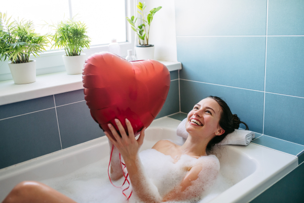 Beautiful woman relaxing in bathtub, holding heart shaped red balloon. Enjoying hot water in bubble bath with music. Valentine's Day and love concept.