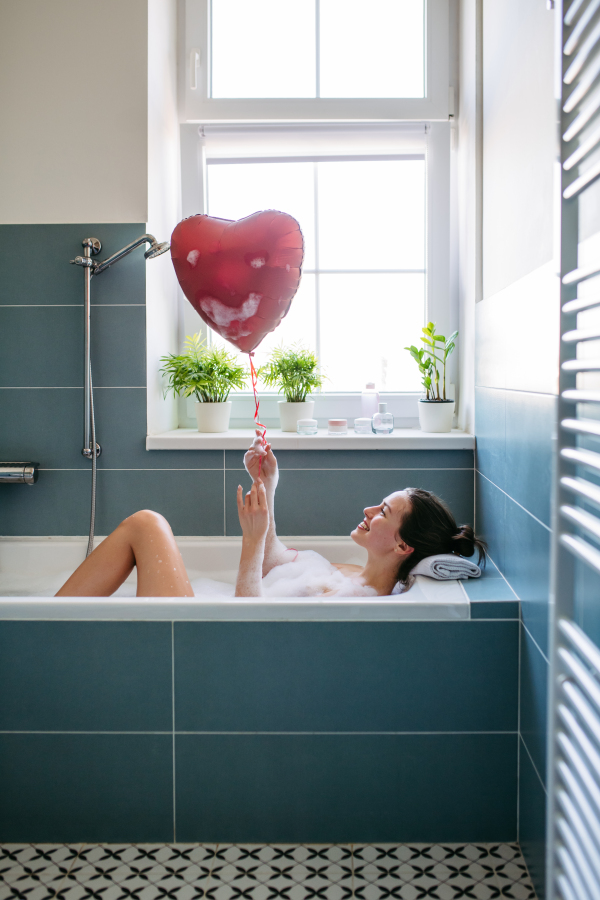 Beautiful woman relaxing in bathtub, holding heart shaped red balloon. Enjoying hot water in bubble bath with music. Valentine's Day and love concept.