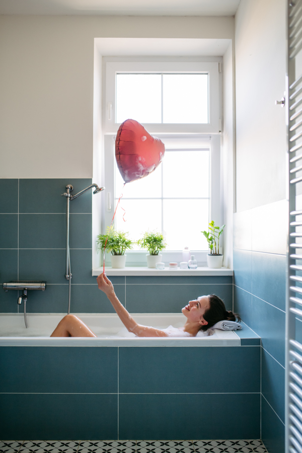 Beautiful woman relaxing in bathtub, holding heart shaped red balloon. Enjoying hot water in bubble bath with music. Valentine's Day and love concept.