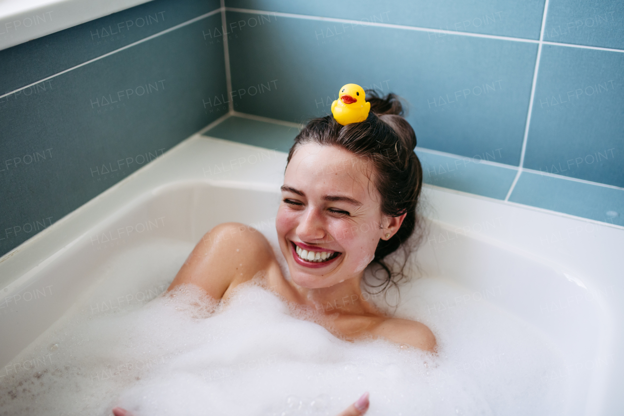 Beautiful young woman in bathtub with rubber duck on head. Enjoying a bubble bath, smiling, having fun.