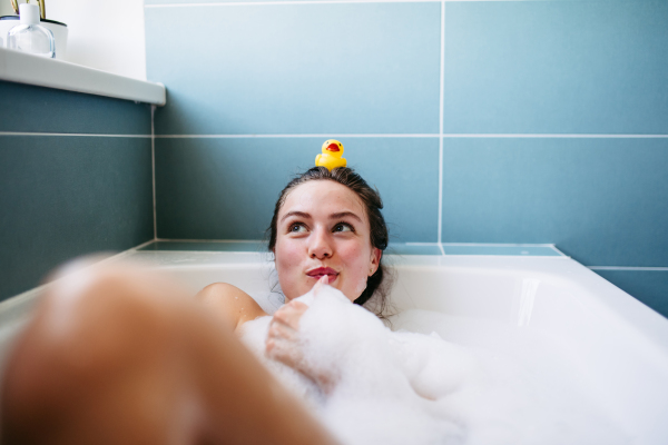 Beautiful young woman in bathtub with rubber duck on head. Enjoying a bubble bath, smiling, having fun.