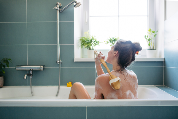 Rear view of woman sitting in warm bathwater, washing her back with bath brush. Bath skincare routine. Soaking in bathtub, enjoying bubble bath.