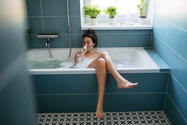 Beautiful woman relaxing in warm bathwater, drinking champagne and soaking in bathtub. Enjoying bubble bath, smiling, looking at camera.