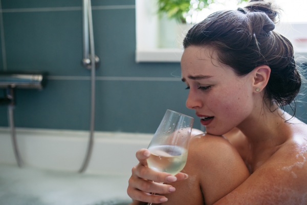 Crying, depressed woman sitting in bathtub, drinking wine. Young woman feeling hopeless, having problems with harassment, rude comments in work.