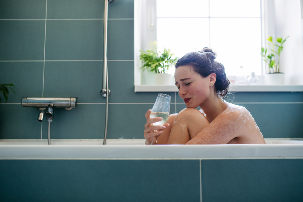 Crying, depressed woman sitting in bathtub, drinking wine. Young woman feeling hopeless, having problems with harassment, rude comments in work.