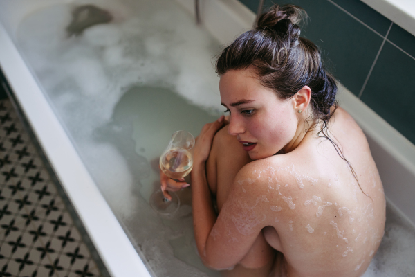 Crying, depressed woman in bathtub, drinking wine. Young woman feeling hopeless, having problems with harassment, rude comments in work.