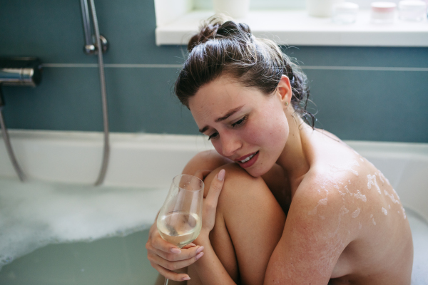 Crying, depressed woman in bathtub, drinking wine. Young woman feeling hopeless, having problems with harassment, rude comments in work.