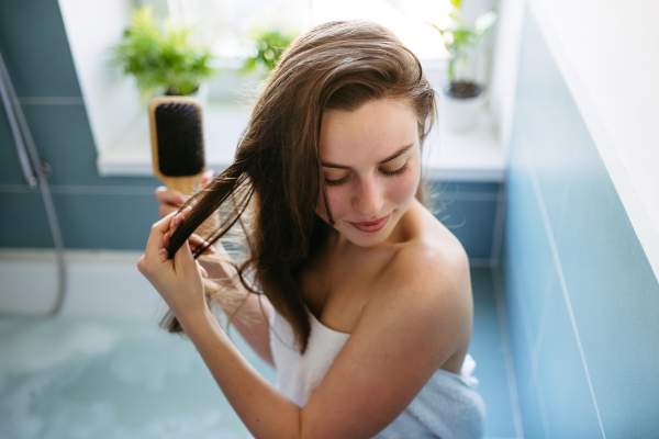 Beautiful young woman wrapped in towel, brushing wet hair in bathroom. Hair care routine for healthy hair.