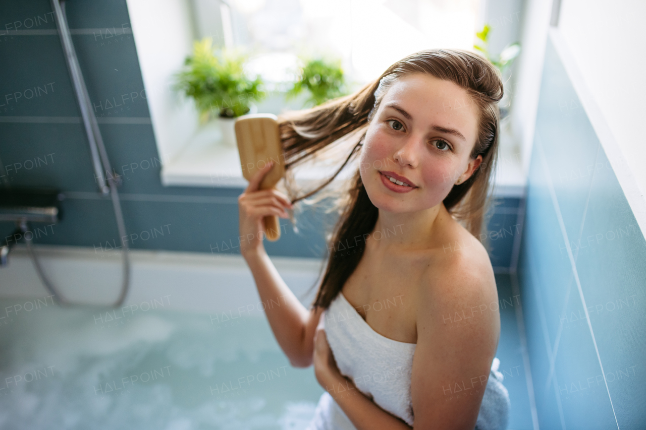 Beautiful young woman wrapped in towel, brushing wet hair in bathroom. Hair care routine for healthy hair.