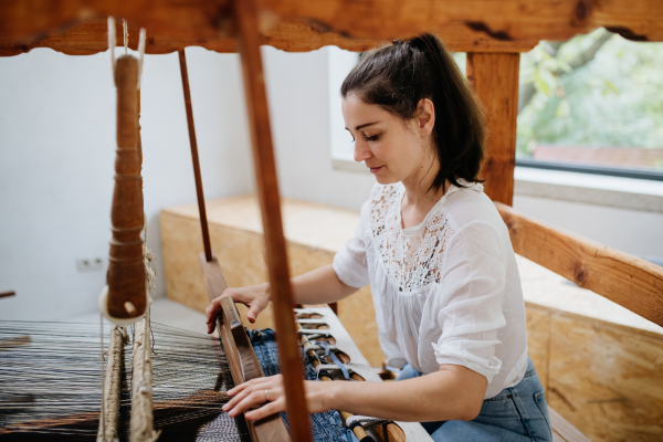 Young businesswoman sitting at a wooden loom, weaving a rug, upholstery from old fabrics. Upcycling in business and circular economy.