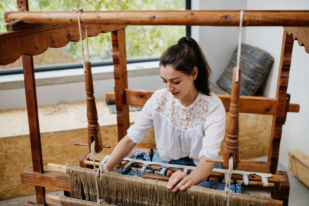 Young businesswoman sitting at a wooden loom, weaving a rug, upholstery from old fabrics. Upcycling in business and circular economy.