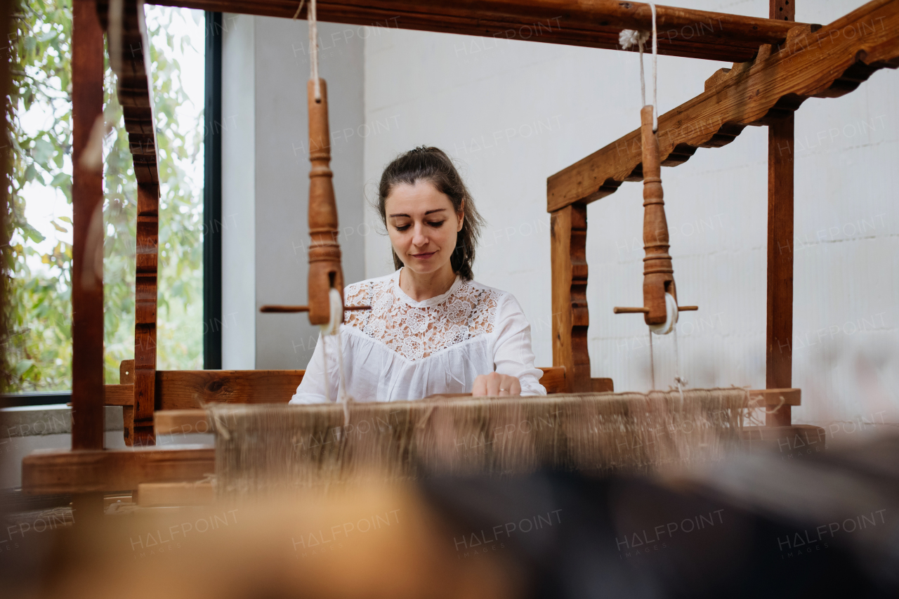 Young businesswoman sitting at a wooden loom, weaving a rug, upholstery from old fabrics. Upcycling in business and circular economy.