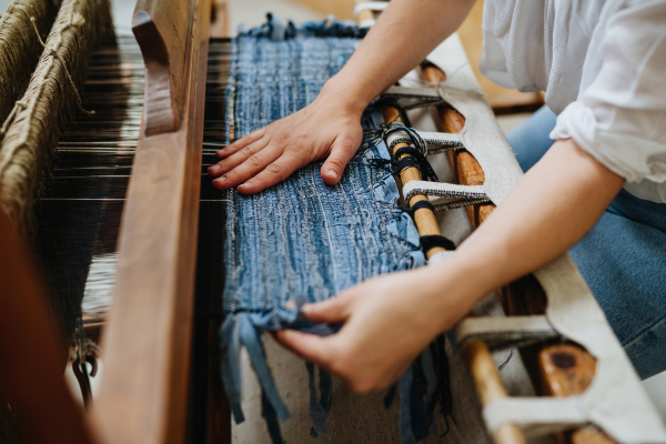 Young businesswoman sitting at a wooden loom, weaving a rug, upholstery from old fabrics. Upcycling in business and circular economy.