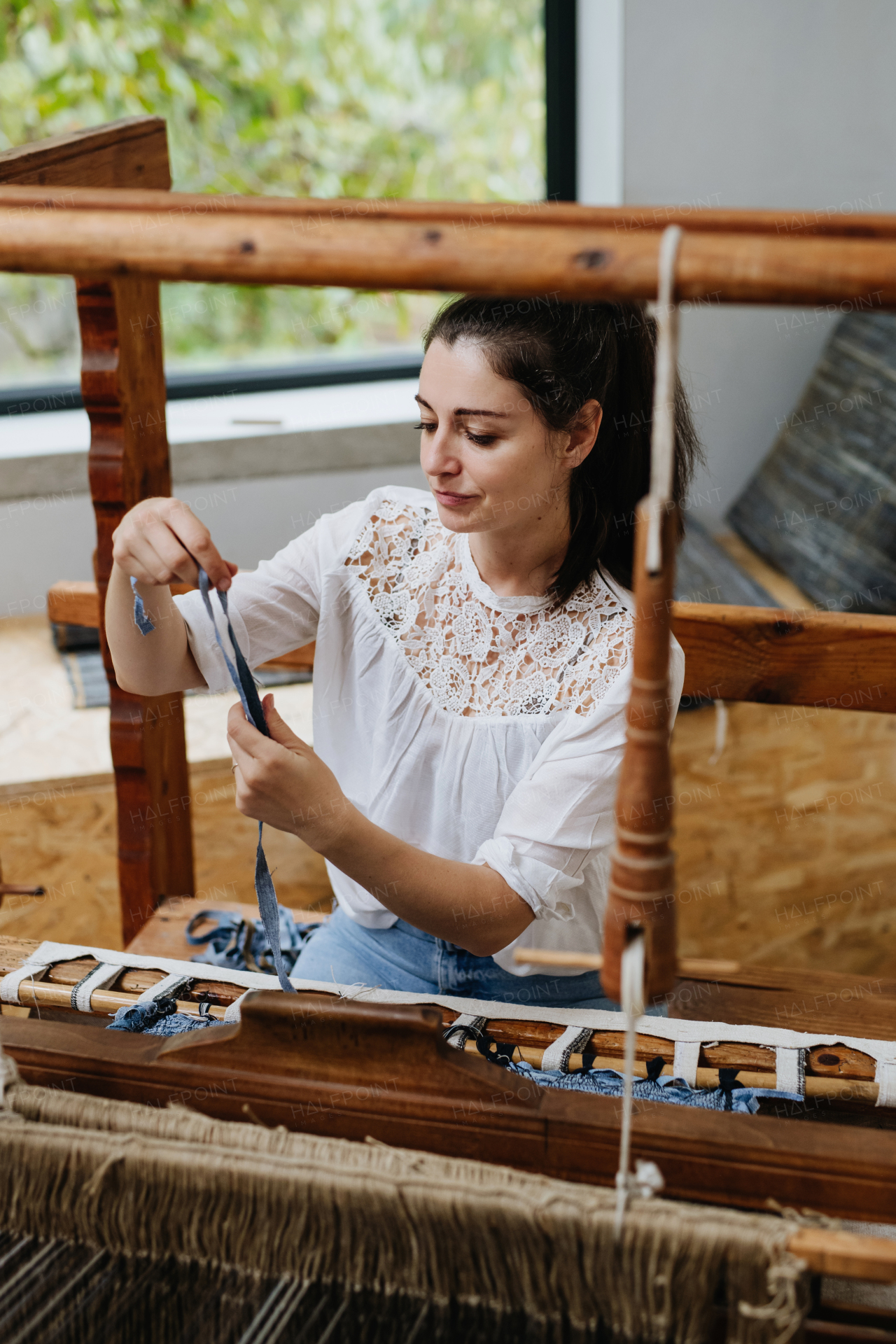 Young businesswoman sitting at a wooden loom, weaving a rug, upholstery from old fabrics. Upcycling in business and circular economy.