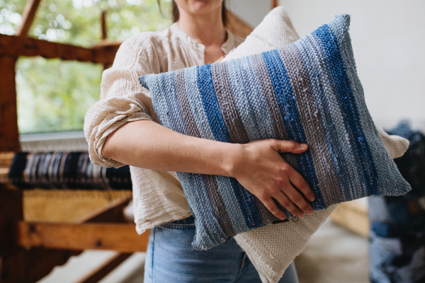 Young businesswoman next to wooden loom, holding pillows with woven covers made from recycled fabrics.. Upcycling in business and circular economy.