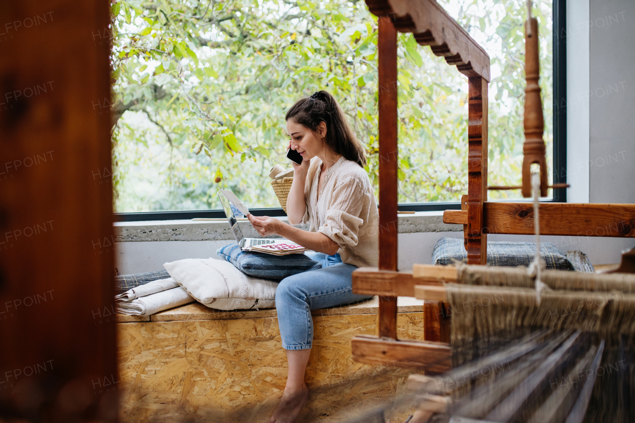 Young businesswoman making a call with customer, sitting by wooden loom. Upcycled textile, small cloth weaving business and circular economy.