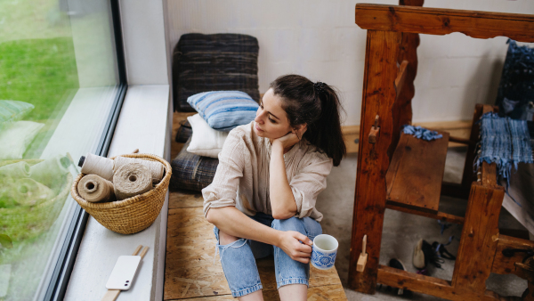 Young businesswoman taking break, sitting next to a wooden loom and drinking coffee. Small weaving business and circular economy.