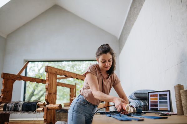 Young businesswoman working on workbench, preparing old fabric for weaving. Upcycled textile, small cloth weaving business and circular economy.