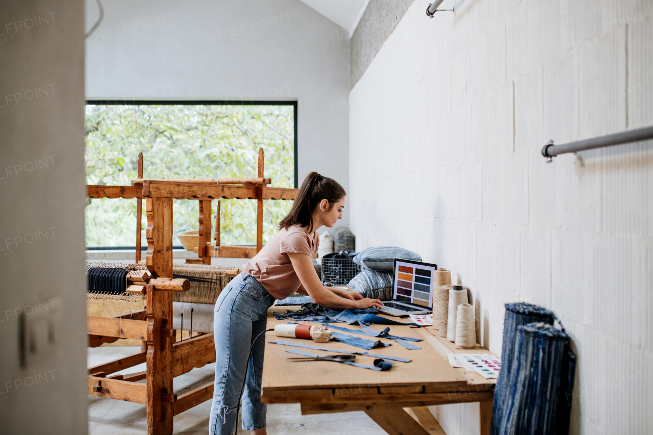 Young businesswoman checking customer order on laptop, standing by wooden loom. Upcycled textile, small cloth weaving business and circular economy.