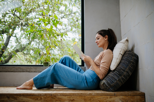 Hygge moment for beautiful woman sitting by large window with a book. Woman is enjoying time for herself, reading.