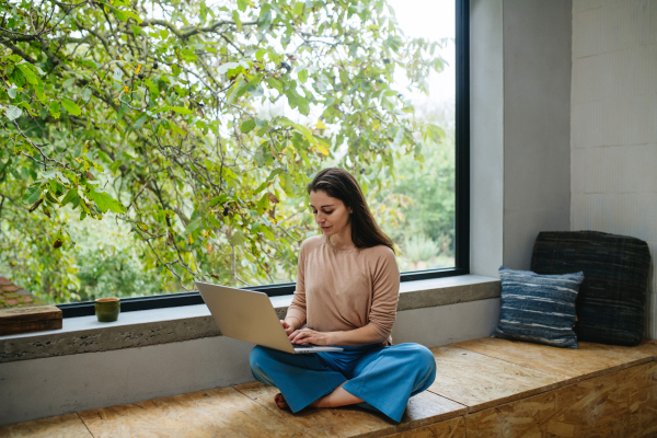 Beautiful woman sitting cross-legged in front of large window, working on laptop.