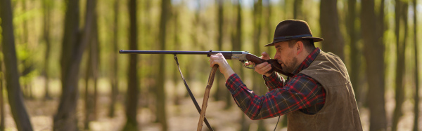 A side view of hunter man aiming with rifle gun on prey in forest.