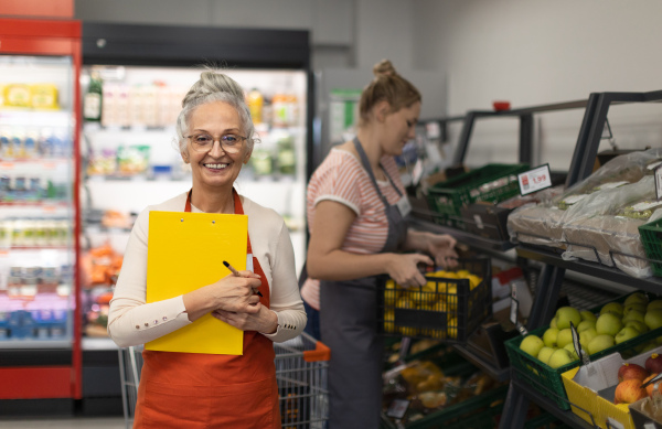 A confident senior shop assistant in supermarket in vegetable shell, in bakcground is her colleague filling stock.