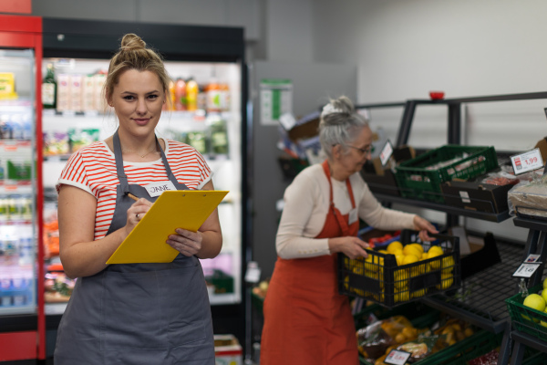 A young shop assistant in supermarket in vegetable shell, in bakcground is her colleague filling stock.