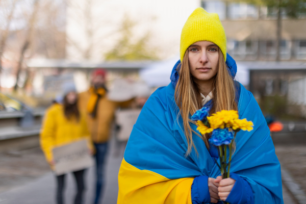 Protest against Russian invasion of Ukraine. A young woman wrapped in Ukrainian flag holding blue and yellow flowers.