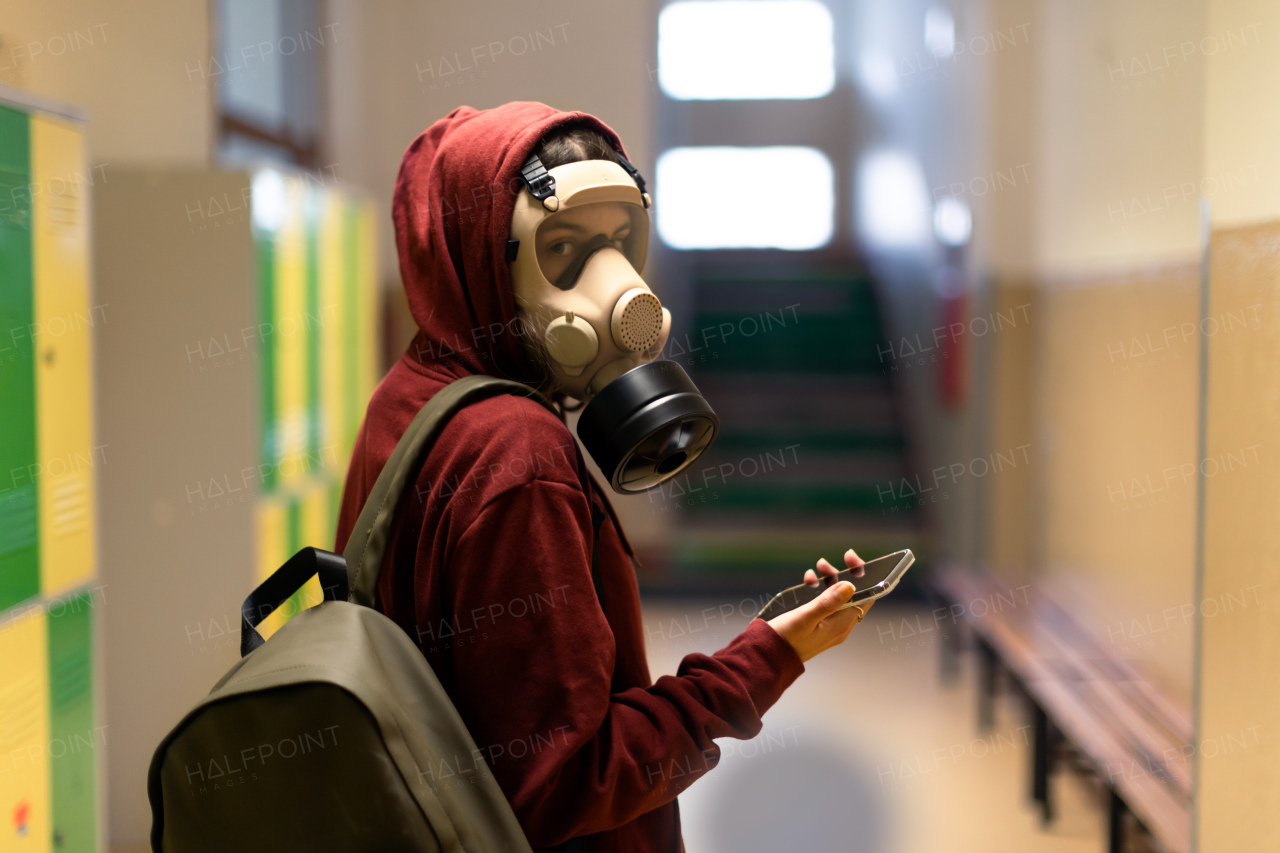 A young student in gas mask with smartphone at school.