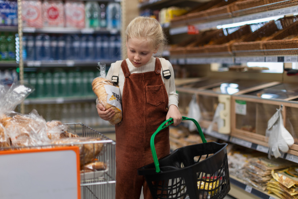 Little girl standing in the middle of supermarket and buying bread, holding shopping basket.