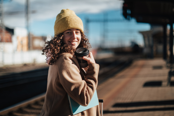 A portrait of happy young traveler woman with coffe and luggage at train station platform