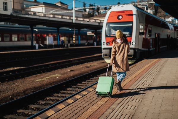 A happy young traveler woman with luggage and cup of coffee boarding in the train at train station platform. Rear view.