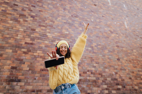 A portrait of happy young woman in front of brick wall in city street, looking at camera.