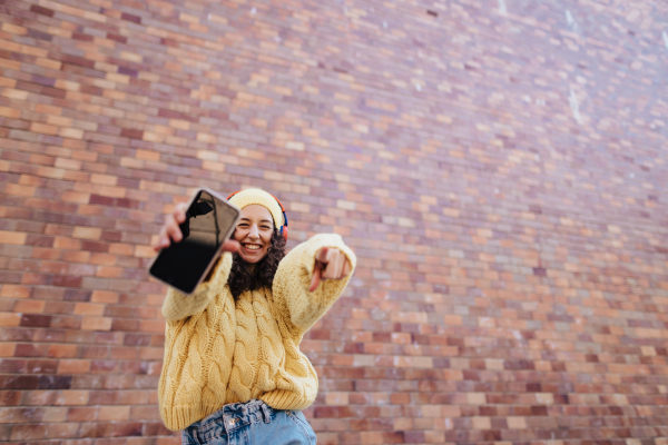 A portrait of happy young woman in front of brick wall in city street, looking at camera.