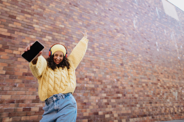 A happy young woman with headphones dancing outdoors in city street, looking at camera.