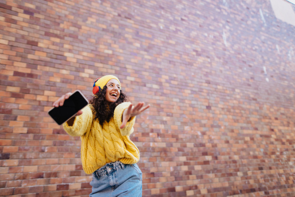 A portrait of happy young woman in front of brick wall in city street, looking at camera.