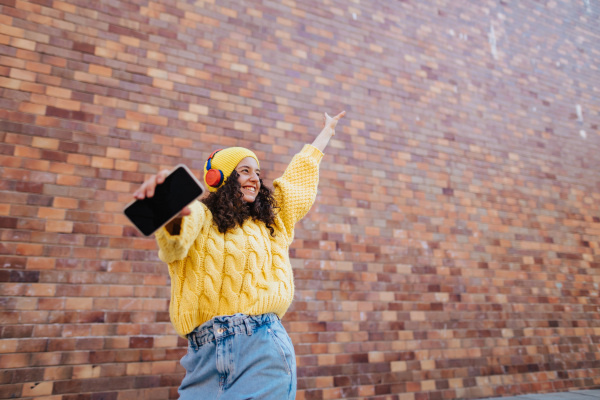 A portrait of happy young woman in front of brick wall in city street, looking at camera.