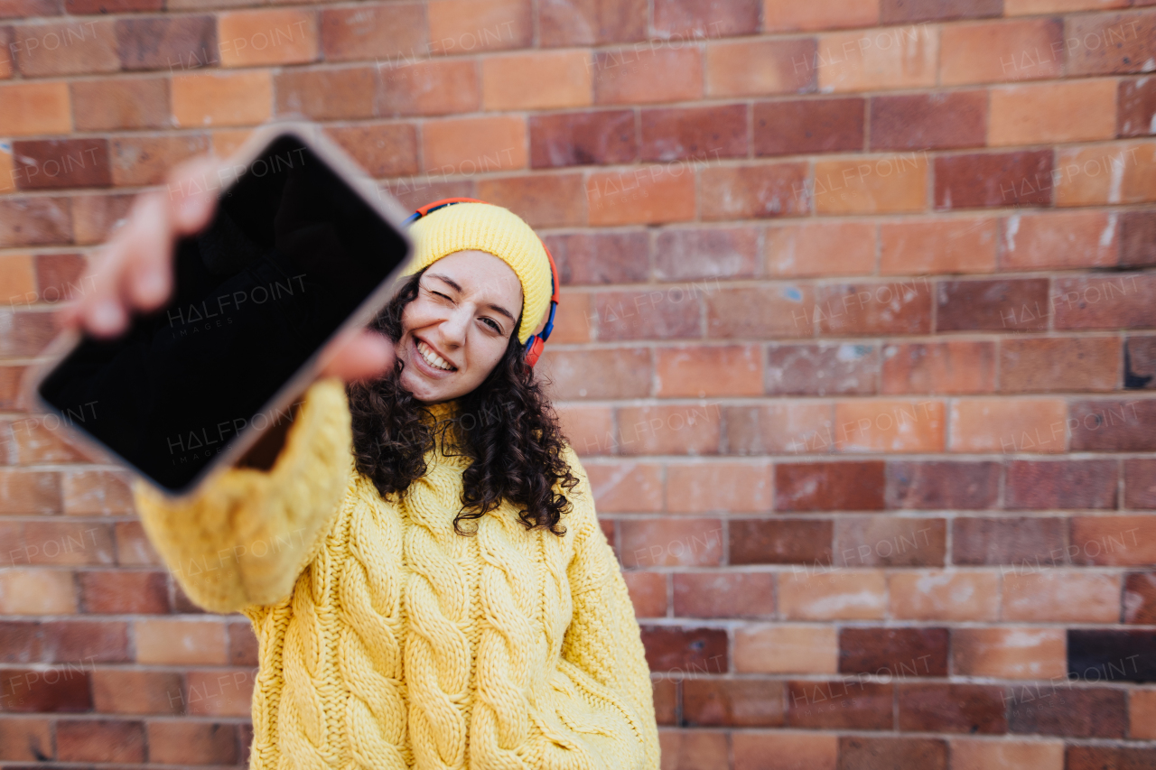 A portrait of happy young woman with smartphone in front of brick wall in city street, looking at camera.