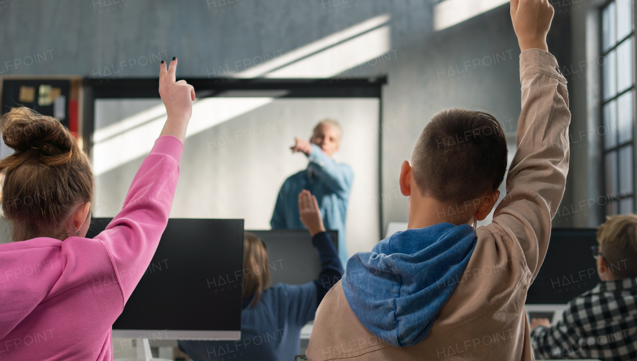 A rear view of school kids raising hands during computer calss at school.