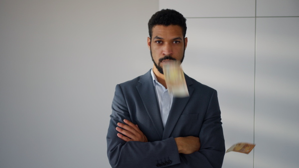 A serious young businessman man standing in office with money falling down around him, inflation concept.